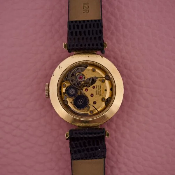 Back view of the Ernest Borel Kaleidoscope Cocktail 17 Jewels gold wristwatch, showing its mechanical gears, displayed on a pink textured surface.