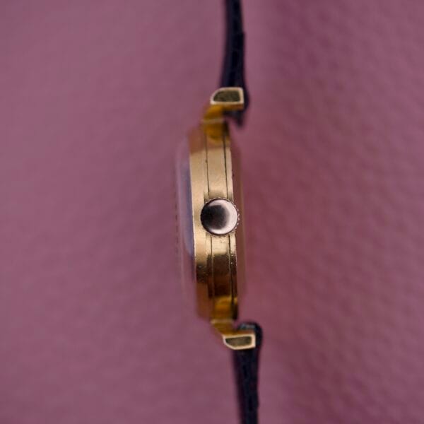 Side view of an Ernest Borel Kaleidoscope Cocktail 17 Jewels gold wristwatch with a black strap, set against a textured pink background.