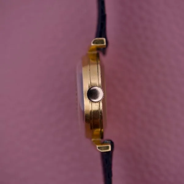 Side view of an Ernest Borel Kaleidoscope Cocktail 17 Jewels gold wristwatch with a black strap, set against a textured pink background.