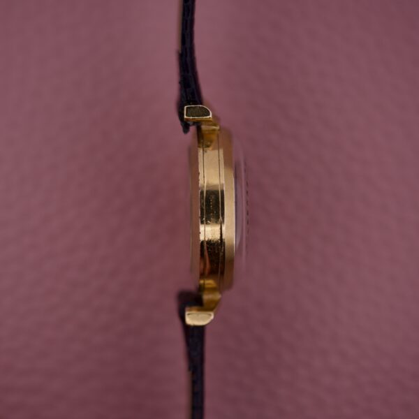 A side view of the Ernest Borel Kaleidoscope Cocktail 17 Jewels gold wristwatch with a black strap, set against a pink textured background.