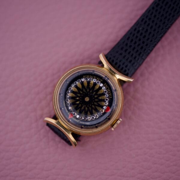The Ernest Borel Kaleidoscope Cocktail 17 Jewels is a gold wristwatch with a black strap on pink textured leather, featuring a unique sunburst dial inspired by the brand’s signature design.
