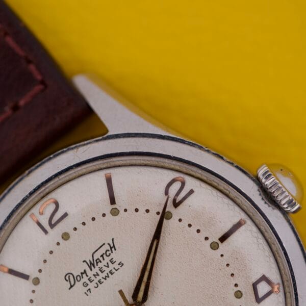 Close-up of a Dom Watch Geneve 17 Jewels Waffle Dial with a brown strap, showing 2:01 on its waffle-patterned face against a yellow background.