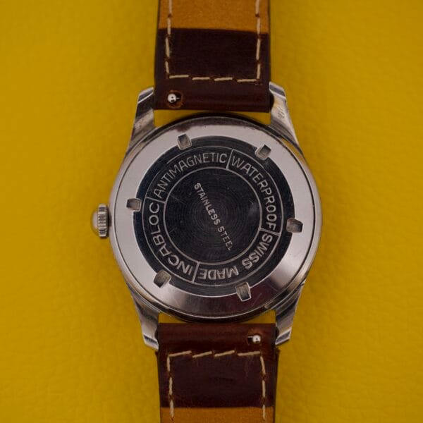 Back of the Dom Watch Geneve 17 Jewels Waffle Dial with brown leather strap on a yellow surface, displaying the engraved Geneve 17 Jewels text and intricate details.