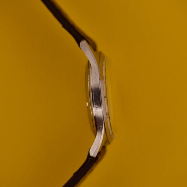 Side view of a silver Dom Watch Geneve 17 Jewels Waffle Dial with a brown strap, set against a solid yellow background.