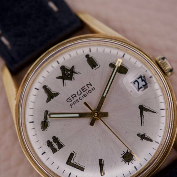 Close-up of a Gruen Precision 510 Date Masonic Dial wristwatch with a gold case, distinctive Masonic symbols marking each hour, and the date shown as 23.