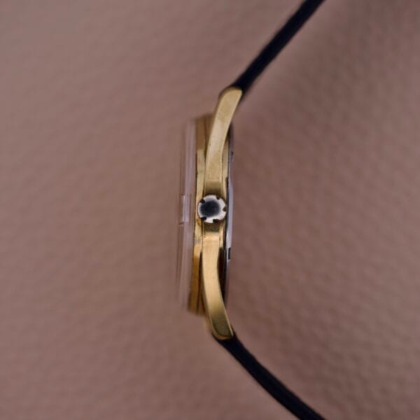 Side view of a Gruen Precision 510 Date Masonic Dial gold wristwatch with black strap and textured crown, shown against a beige background.