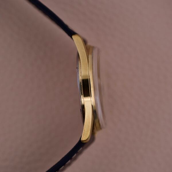 Side view of the Gruen Precision 510 Date Masonic Dial gold wristwatch case with black strap, shown against a textured pink background.