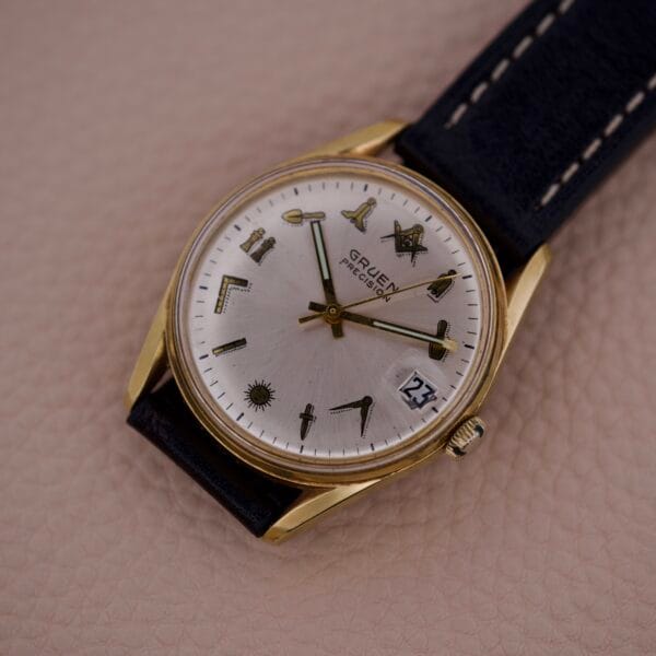 A Gruen Precision 510 Date Masonic Dial wristwatch featuring a gold case, black leather strap, silver Masonic dial, and distinctive symbol hour markers.