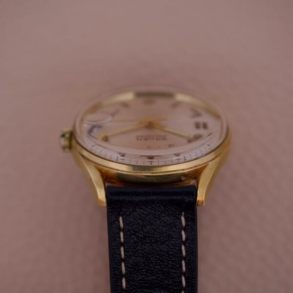 The Gruen Precision 510 Date Masonic Dial wristwatch features a gold case, black leather strap, and Masonic dial, displayed from the side against a beige background.