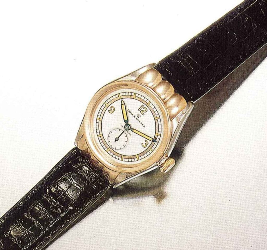 A vintage Rolex Oyster Perpetual wristwatch with a black leather strap and a round gold case is displayed on a white background.