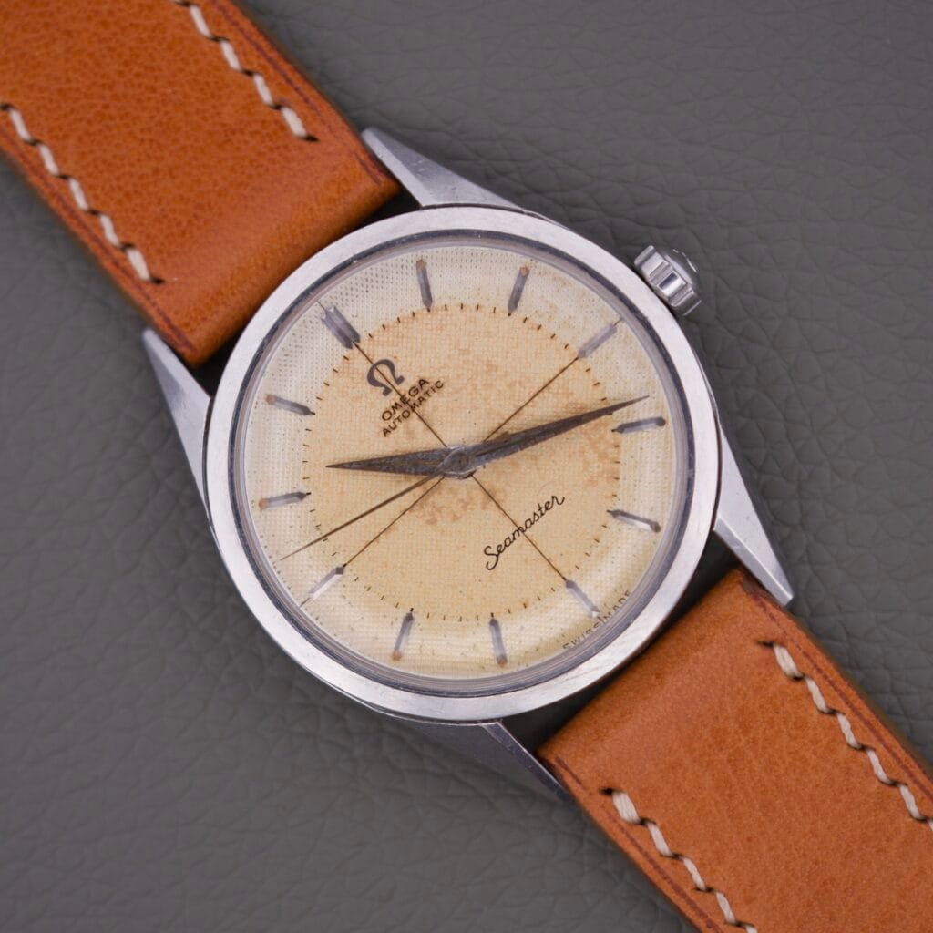 Omega Automatic Seamaster 2802 Cross Hair Waffle Dial watch with a classic tan leather strap is displayed on a gray textured surface.