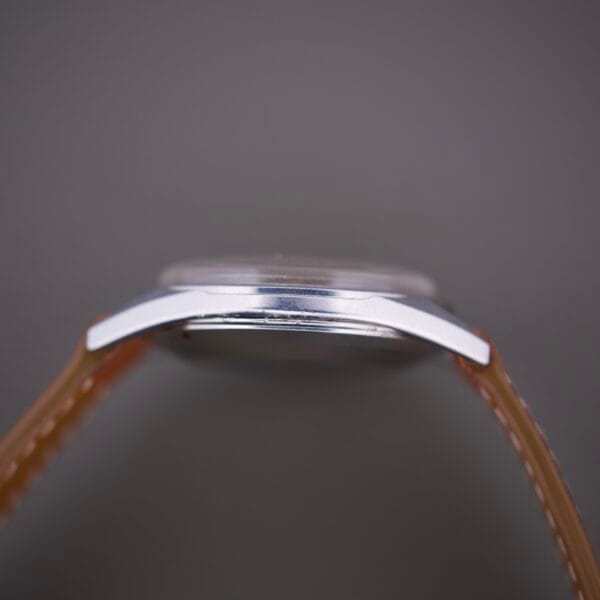 Side view of an Omega Automatic Seamaster 2802 Cross Hair Waffle Dial wristwatch with a brown strap and automatic movement, set against a gray background.