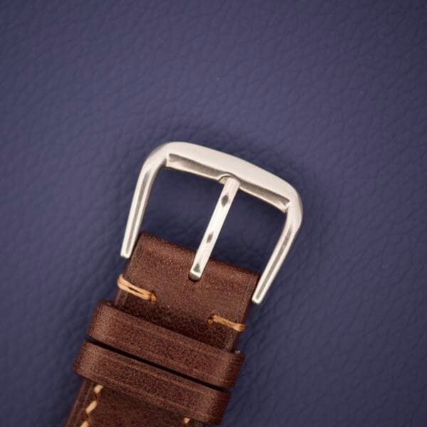 Close-up of a brown leather watch strap with a silver buckle on a dark blue textured background, capturing the timeless elegance of the Rolex Datejust 1603 Silver Dial Alpha hands.