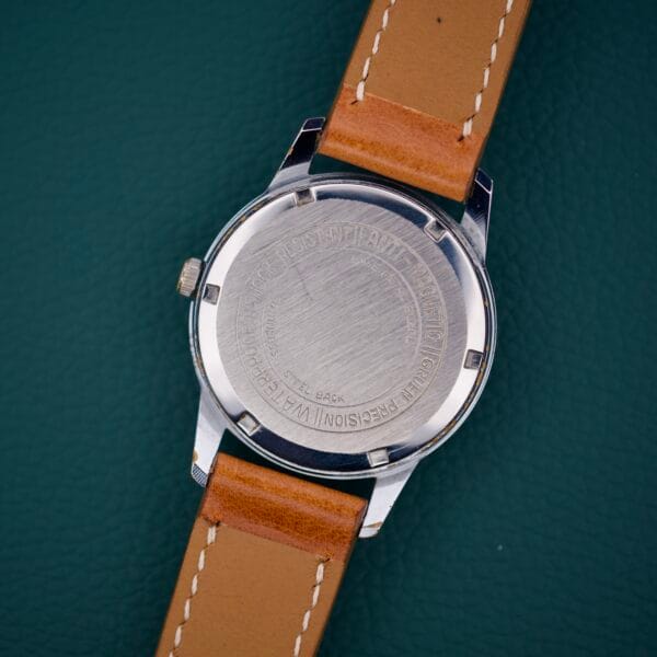 A Gruen Precision 510 RSS Day / Night Jump Hour Markers wristwatch with a tan leather strap is shown face-down, revealing its stainless steel back on a green surface.