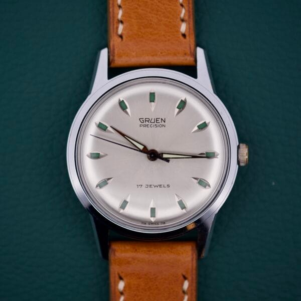 Gruen Precision 510 RSS Day / Night Jump Hour Markers wristwatch with a brown leather strap displayed on a dark green textured background.