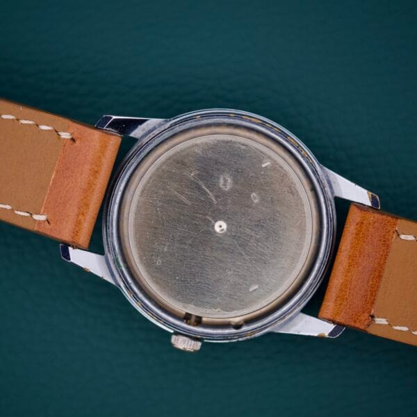 The Gruen Precision 510 RSS Day / Night Jump Hour Markers wristwatch, featuring a blank face and brown leather strap, is displayed against a dark green textured background.