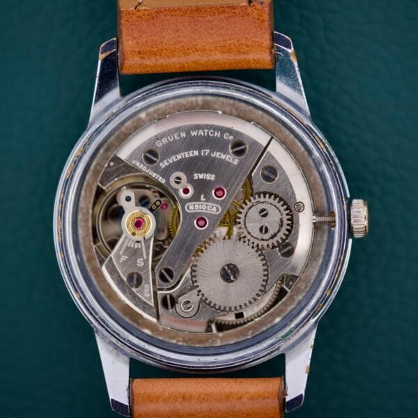 A close-up of the Gruen Precision 510 RSS Day / Night Jump Hour Markers watch’s gears and parts, featuring a brown leather strap against a green background.