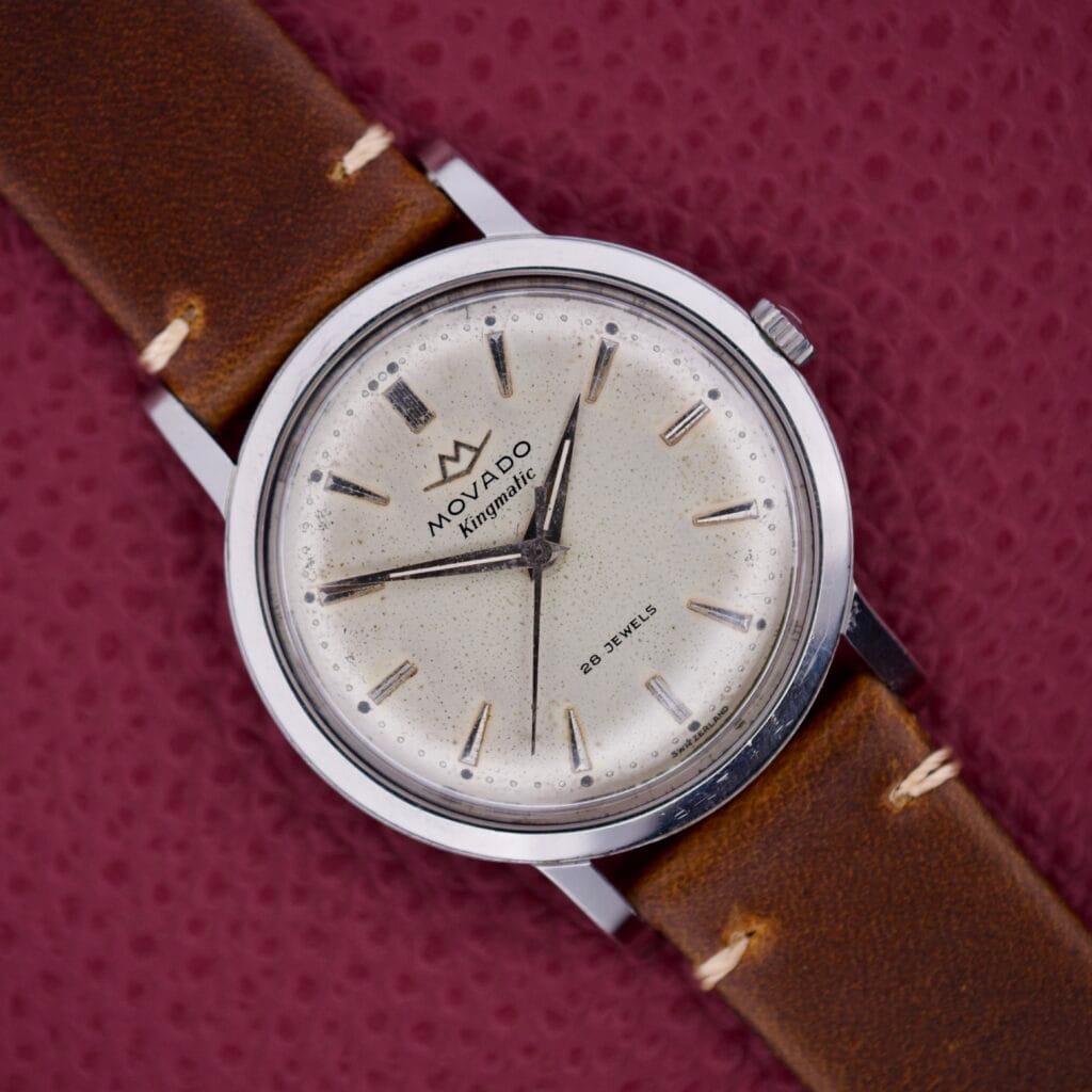 Movado Kingmatic Ref. 15051 28 Jewels wristwatch with a brown leather strap displayed on a textured burgundy surface.