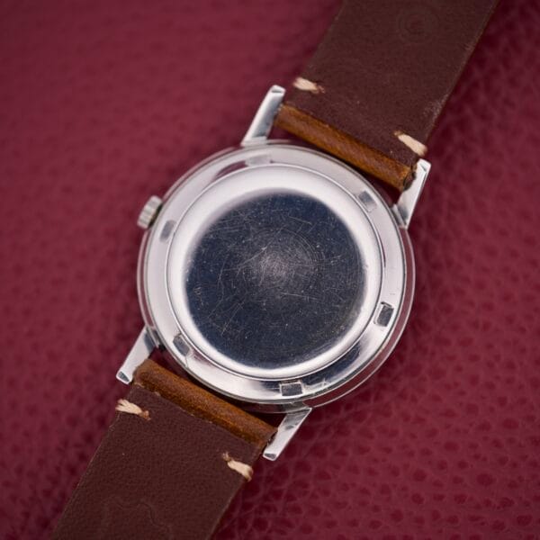 A Movado Kingmatic Ref. 15051 28 Jewels wristwatch with a brown leather strap and scratched metal case back rests on a red textured surface.