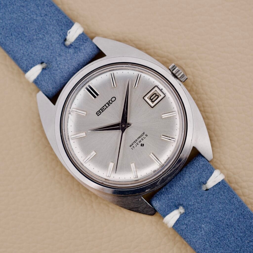 Seiko 6602-8040 Manual Wind Date Sunburst Dial wristwatch with a blue strap and date display shown on a tan surface.