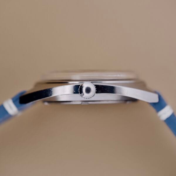 Side view of the Seiko 6602-8040 Manual Wind Date Sunburst Dial wristwatch in silver with a blue strap, displayed against a beige background.