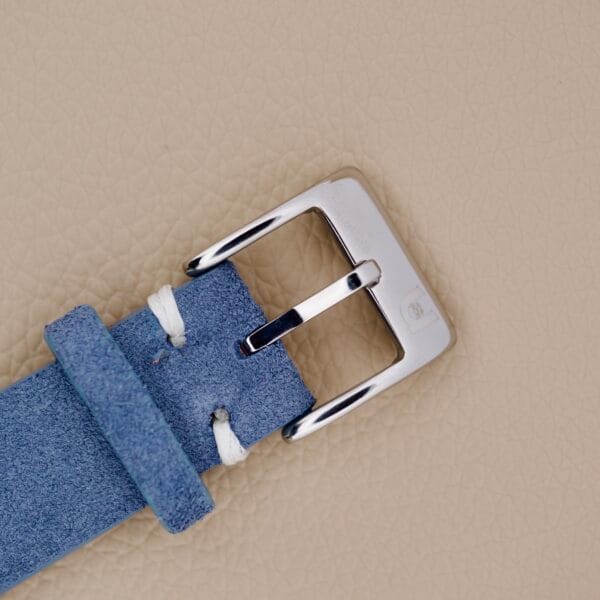 Close-up of a blue suede belt with a silver buckle on a beige textured background, evoking the refined style of the Seiko 6602-8040 Manual Wind Date Sunburst Dial.