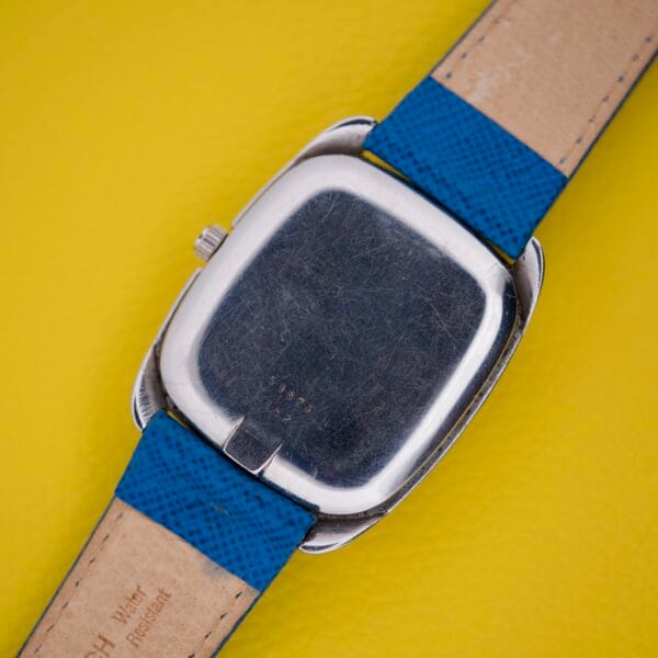 The Favre Leuba Tornado Blue Dial Automatic Date silver wristwatch, with blue and tan straps, is displayed face down on a yellow textured background.