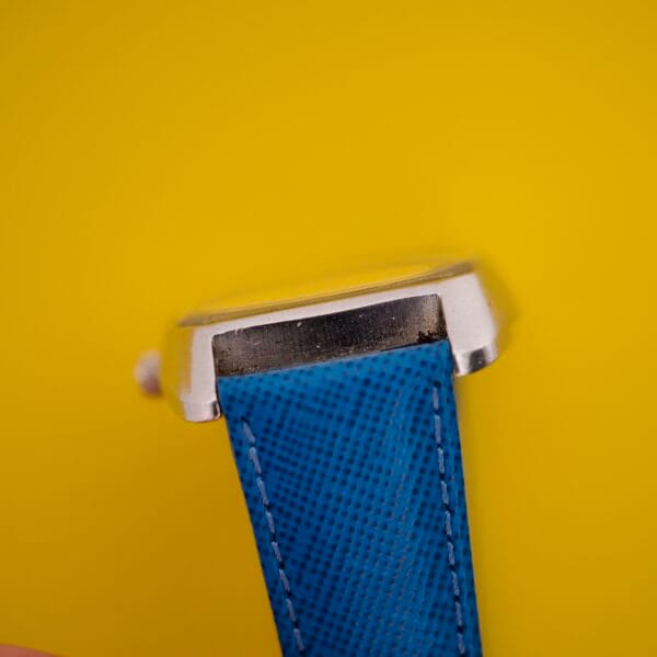 Close-up of a Favre Leuba Tornado Blue Dial Automatic Date silver wristwatch case with a blue textured strap, set against a yellow background.