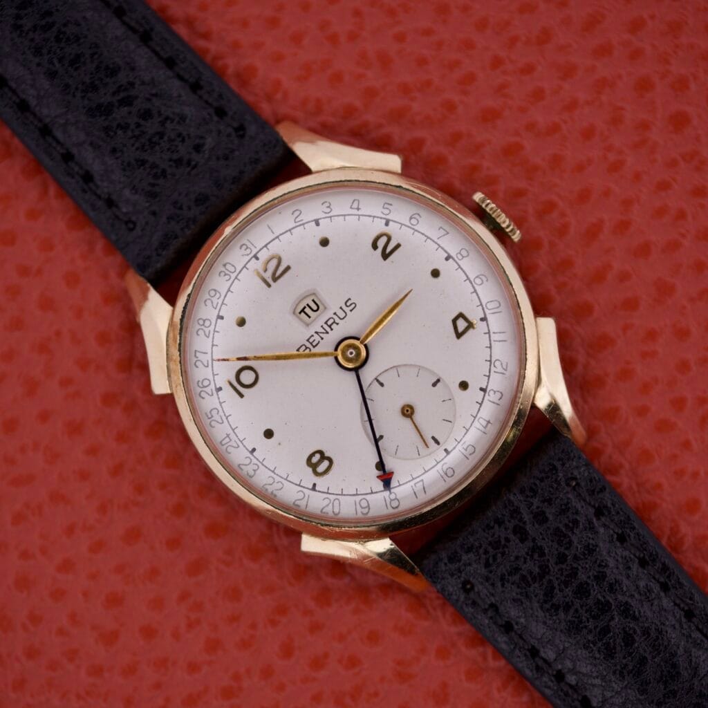 A Benrus Triple Calendar Pointer Model CE 13 gold wristwatch with a black leather strap rests on a textured red surface.