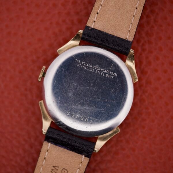 Back of a Benrus Triple Calendar Pointer Model CE 13 wristwatch with leather strap, showing engraved text, serial number, and CE 13 on a red surface.