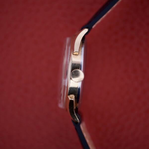 Side view of a Benrus Triple Calendar Pointer Model CE 13 gold wristwatch with a black strap, displayed against a textured red background.