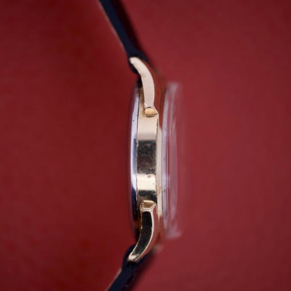 Side view of the Benrus Triple Calendar Pointer Model CE 13 gold wristwatch with a black strap, highlighting its classic calendar feature against a striking red background.