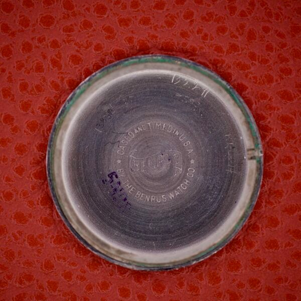 A round metal case back from the Benrus Triple Calendar Pointer Model CE 13, stamped with text, displayed on a red textured surface.