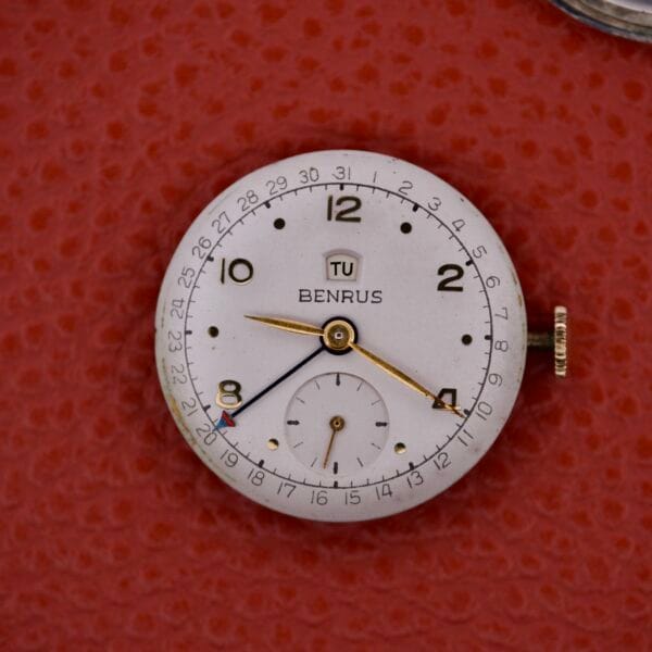 A Benrus Triple Calendar Pointer Model CE 13 watch rests elegantly on a vibrant red surface.