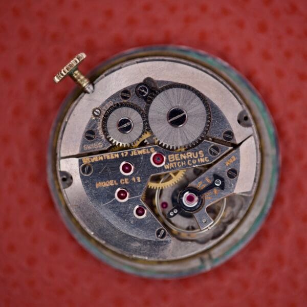Close-up of the intricate gears and mechanism of the Benrus Triple Calendar Pointer Model CE 13, shown against a red textured background.