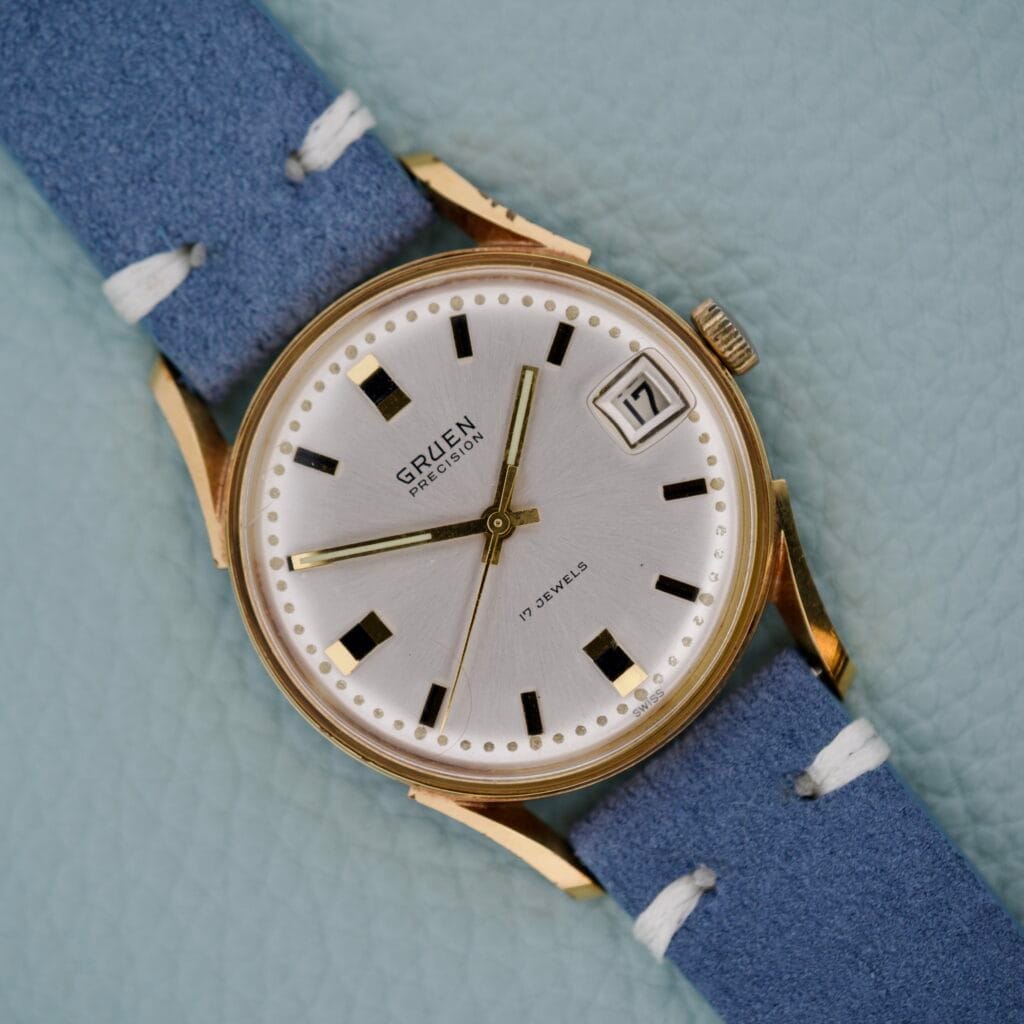 Gruen Precision Date 510RSS CA wristwatch with gold case, blue strap, and sunburst dial, shown on a light blue background.