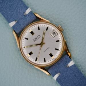 Gruen Precision Date 510RSS CA wristwatch with gold case, blue strap, and sunburst dial, shown on a light blue background.