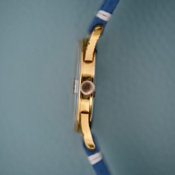 Side view of the Gruen Precision Date 510RSS CA Sunburst Dial gold wristwatch with a blue strap, displayed against a textured light blue background.