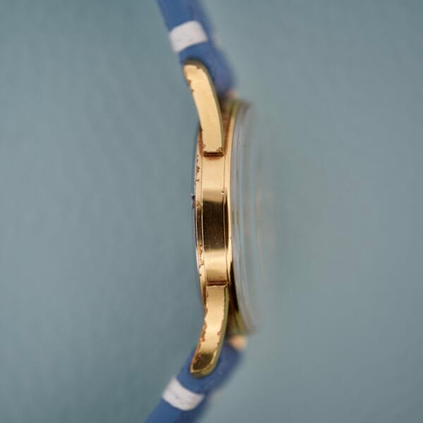 Side view of the Gruen Precision Date 510RSS CA Sunburst Dial gold wristwatch case with sunburst dial and blue strap, set against a light blue textured background.