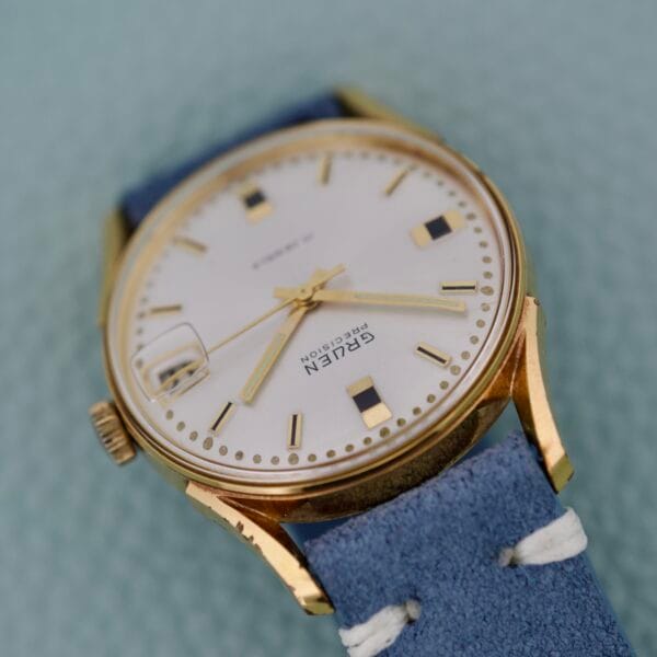 Gruen Precision Date 510RSS CA Sunburst Dial wristwatch with a blue leather strap displayed on a textured light green background.