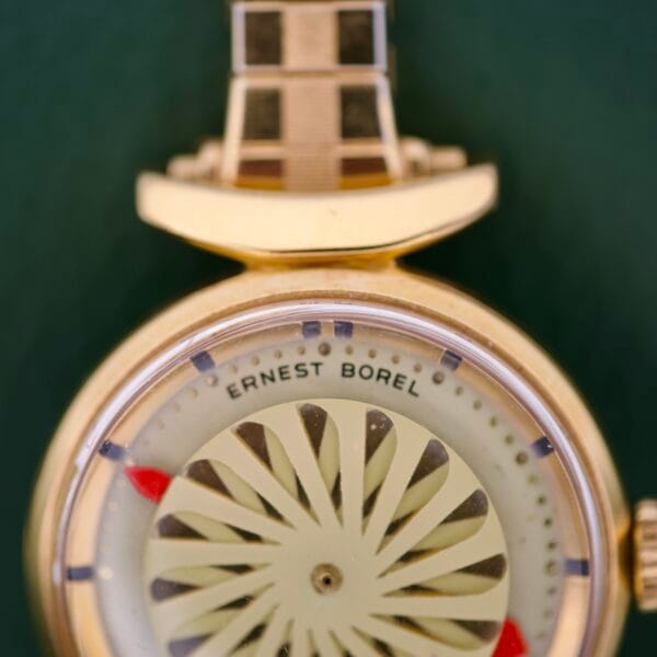 Close-up of an Ernest Borel Cocktail Kaleidoscope White Dial wristwatch with a patterned face and red arrow markers, set against a green background.