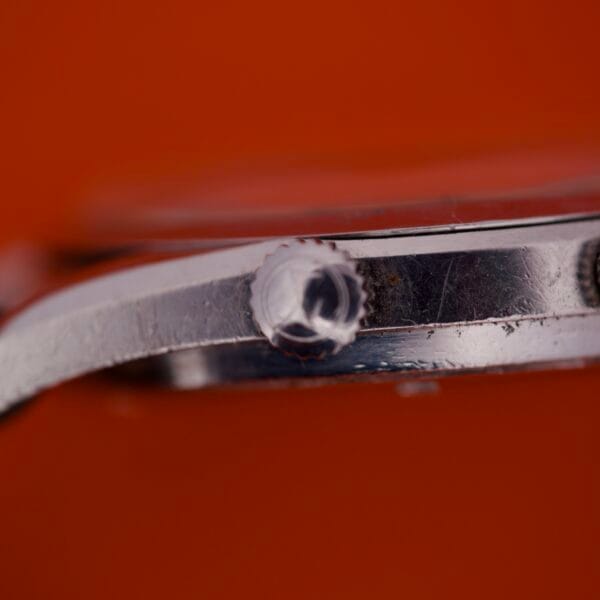 Close-up of the Favre Leuba Geneve Seabird Alarm Stainless Steel watch crown and case against a solid orange background.