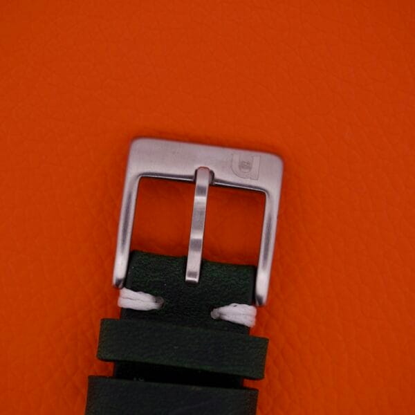 A close-up of a Favre Leuba Geneve Seabird Alarm Stainless Steel buckle on a dark leather strap, set against an orange textured background.