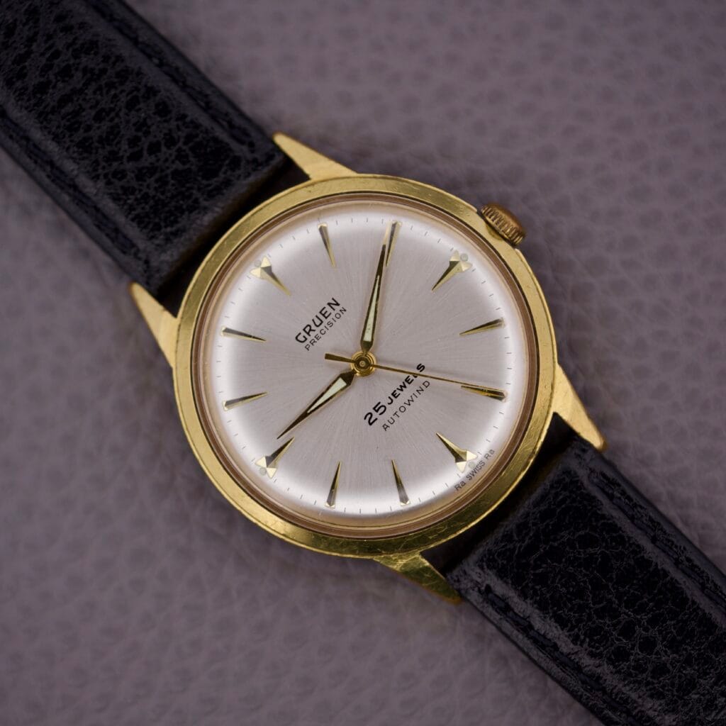 The Gruen Precision 25 Jewels Autowind Sunburst Dial wristwatch with a black leather strap is elegantly displayed on a textured grey surface.