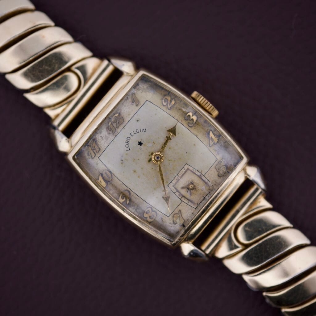 A vintage Lord Elgin Art Deco Caliber 559 21 Jewels USA wristwatch with a rectangular face and flexible metal band rests on a dark surface.