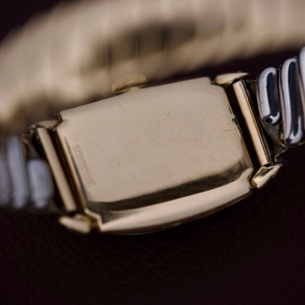 Close-up of the back of a Lord Elgin Art Deco Caliber 559 21 Jewels USA gold wristwatch with a metal band, highlighting classic Art Deco details on a dark textured surface.