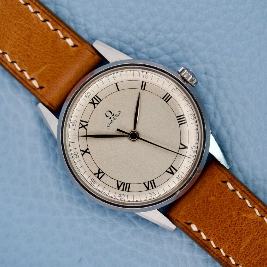 An Omega Ref. 2271-2 Jumbo stainless steel wristwatch with a tan leather strap and Roman numeral dial, powered by the 30T2 movement, is displayed elegantly on a light blue textured surface.