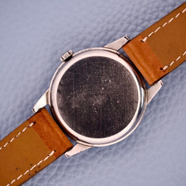 An Omega Ref. 2271-2 Jumbo Stainless Steel Case 30T2 wristwatch with a scratched metal back and brown leather strap rests on a textured light blue surface, highlighting its vintage design.