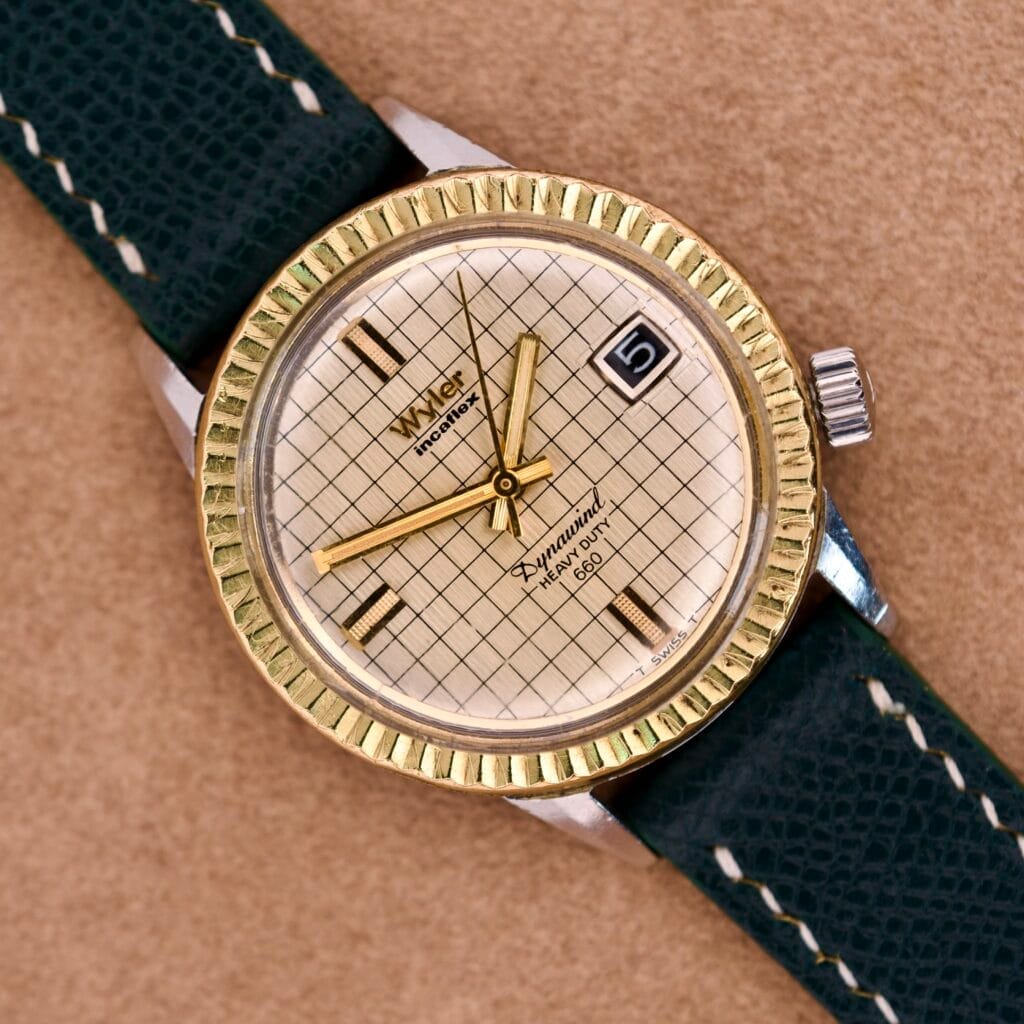 A Wyler Dynawind Heavy Duty 660 Life Guard wristwatch with a grid-patterned face, gold accents, and a dark leather strap rests on a tan surface.