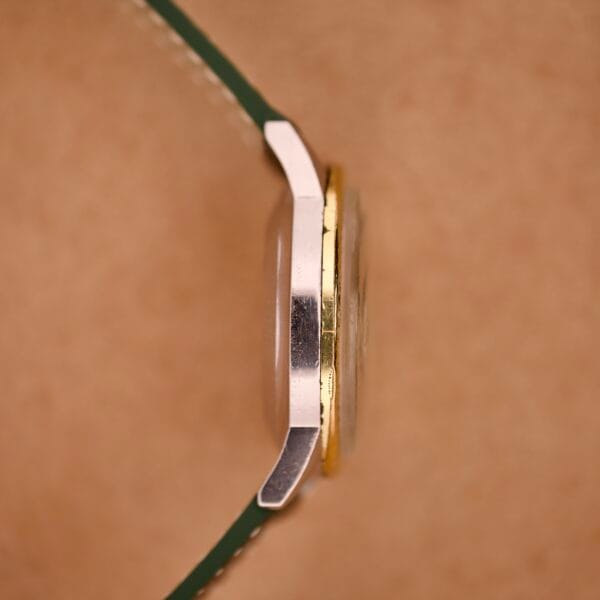 Side view of a Wyler Dynawind Heavy Duty 660 Life Guard wristwatch with a green strap and gold trim, set against a soft brown background.